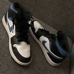 Nike Air Jordan 1 Mid svart/blå/vit - Säljer ett par Nike Air Jordan 1 Mid i svart, blått och vitt. Skorna har klassisk hög siluett, svart snörning och vit sula. Perfekta för dig som gillar streetwear och vill sticka ut med snygga sneakers.