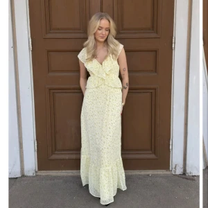 Ljusgul långklänning med volang och blommigt mönster - Söt ljusgul långklänning med små blommor och volangdetaljer framtill. Klänningen har v-ringning och är ärmlös, perfekt för sommarens alla tillfällen. Bekväm passform och romantisk stil. Denna klänningen är aldrig använd, testad en gång.