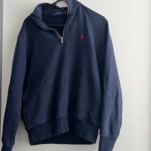Mörkblå halvzip tröja från Polo Ralph Lauren - Säljer en mörkblå långärmad halvzip tröja från Polo Ralph Lauren med klassisk röd logga broderad på bröstet. Tröjan har hög krage med dragkedja och ribbade muddar vid ärmslut och nederkant.