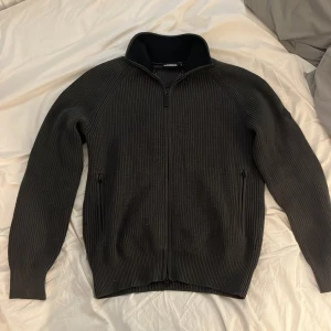 Väldigt mörkgrön stickad kofta från J.Lindeberg - Säljer en väldigt mörkgrön stickad kofta från J.Lindeberg med hel dragkedja framtill och hög krage. Tröjan har långa ärmar och ribbad struktur, samt två fickor med dragkedja.