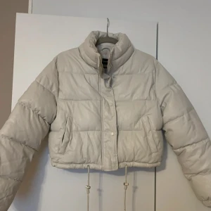  croppad pufferjacka - Snygg beige/ljusgrå pufferjacka med croppad passform och hög krage. Jackan har långa ärmar, dragkedja och knappar framtill samt justerbar dragsko i nederkant.