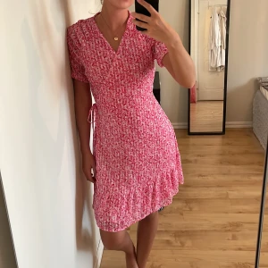 Rosa blommig omlottklänning - Söt rosa omlottklänning med blommigt mönster och volangdetalj nertill. Klänningen har korta ärmar, v-ringning och knyt i midjan för en smickrande passform. Perfekt för varma dagar och somriga tillfällen. Precis som ny❤️