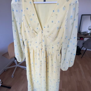 Gul blommig klänning  - Söt gul långklänning med små blå blommor och v-ringning. Klänningen har långa, transparenta ärmar och dekorativa tygklädda knappar framtill. Perfekt för vår och sommar. Den har brustit lite i en sömn (se sista bilden) men inget som syns! 