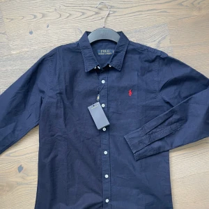 Mörkblå skjorta från Polo Ralph Lauren - Klassisk mörkblå skjorta från Polo Ralph Lauren med knappar framtill och röd broderad logga på bröstet. Skjortan har lång ärm och stilren krage, perfekt för en snygg och enkel look.