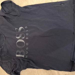 PP/Boss -T-shirt - Svart t-shirt från Hugo Boss med stor glittrig BOSS-logga framtill i silver. Klassisk passform och rund halsringning. Perfekt för dig som gillar stilrena och snygga plagg med lite extra detalj.