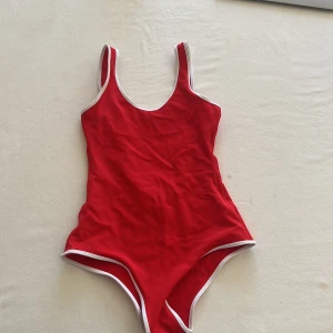 Röd baddräkt från Nayomi - Snygg röd baddräkt från Nayomi med vita kanter och klassisk skärning. Baddräkten har djup rygg och är tillverkad i ett stretchigt material som passar perfekt för strand eller pool. Aldrig använd
