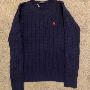 Mörkblå kabelstickad tröja från Polo Ralph Lauren - Säljer en klassisk mörkblå kabelstickad tröja från Polo Ralph Lauren med rund halsringning och den ikoniska röda loggan broderad på bröstet. Tröjan har långa ärmar och ribbade muddar vid ärmslut och nederkant. Kan sänka vid snabba affära