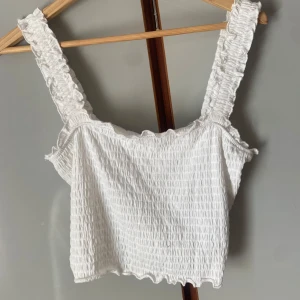 Vit smockad linnetopp från Vero Moda - Superfin vit linnetopp från Vero Moda med smockat tyg och breda axelband. Toppen är croppad och har en söt volangkant både upptill och nertill. Perfekt till sommaren! Lapparna kvar