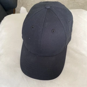 Svart keps från Take Off - En stilren svart keps från Take Off med klassisk böjd skärm och justerbar rem baktill. Kepsen är tillverkad i bomull och har ventilationshål för extra komfort. Perfekt för en enkel och clean look.