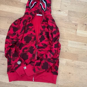 Röd camouflage hoodie från Bape - Säljer en röd camouflage hoodie från Bape med ikoniskt hajmotiv på huvan och broderade detaljer. Jackan har dragkedja hela vägen upp, ribbade muddar och klassisk Bape-logga på ärmen.