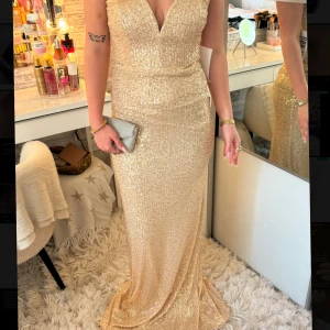 Guldig glittrig långklänning med v-ringning - Säljer en elegant guldig långklänning med djup v-ringning och glittrigt material. Klänningen har figurnära passform och är ärmlös, perfekt för speciella tillfällen. Bärs med en liten clutch för extra glamour.