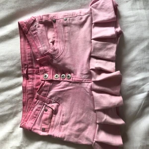 Rosa minikjol med volang och knappar - Säljer en söt rosa minikjol med volangkant och dekorativa knappar framtill. Kjolen har klassiska jeansfickor både fram och bak samt bälteshällor. Perfekt för dig som vill sticka ut med en lekfull och trendig stil!