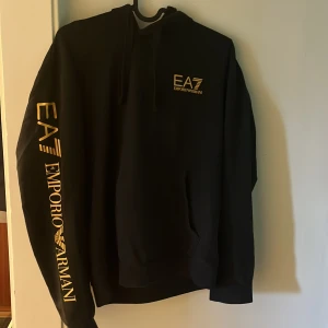Svart hoodie från Emporio Armani EA7 - Säljer en svart hoodie från Emporio Armani EA7 med guldigt tryck på bröstet och längs ena ärmen. Tröjan har huva och ribbade muddar. Perfekt för en sportig och stilren look.