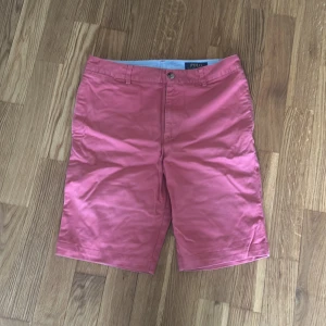 Rosa shorts från Polo Ralph Lauren - Snygga rosa shorts från Polo Ralph Lauren med klassisk passform. Shortsen har två bakfickor med knappar och stängs med dragkedja och knapp framtill. Perfekta för sommaren och tillverkade i bomull.