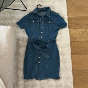 Jeansklänning med bälte och kort ärm - Snygg jeansklänning i blå denim med korta ärmar, knappar framtill och tillhörande bälte i midjan. Klänningen har två bröstfickor med lock och klassisk krage. Perfekt för en trendig och avslappnad look.