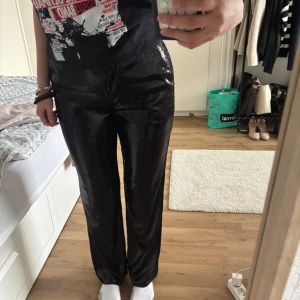 Svarta glittriga byxor - Säljer ett par svarta glittriga byxor med rak passform. Byxorna har hög midja och är perfekta för dig som vill sticka ut med en cool look. Materialet är glansigt och reflekterar ljuset snyggt.