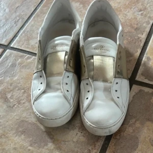 Valentino Open sneakers i vitt och guld - Säljer ett par Valentino Open sneakers i vitt skinn med metallicguld detalj över vristen. Klassisk rund tå och platt sula. Märkeslogga på plösen och sula. Perfekta för dig som gillar lyxiga och trendiga sneakers.