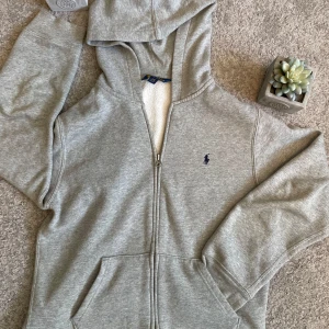 Grå Ralph Lauren Zipp hoodie - Hej! Säljer min grå Ralph lauren Zipp upp hoodie med broderad logga på bröstet. Tröjan har huva, fickor framtill och är tillverkad i mjuk bomull. Hoodien är i mycket bra skick och har knappt använts. Storleken är L/G 14-16 160. Om du har några funderingar, tveka inte om att höra av dig! 😊