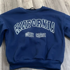Mörkblå sweatshirt med tryck  - Säljer en mörkblå sweatshirt med vitt tryck 'California West Coast' på bröstet. Tröjan har rund halsringning, långa ärmar och ribbade muddar vid ärmslut och nederkant. Perfekt för en avslappnad stil.