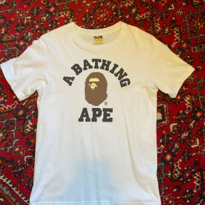 Vit t-shirt från A Bathing Ape - Säljer en vit t-shirt från A Bathing Ape med det klassiska apansikte-trycket och texten 'A BATHING APE' på bröstet. T-shirten är i 100% bomull och har en normal passform med korta ärmar. Perfekt för dig som gillar streetwear!
