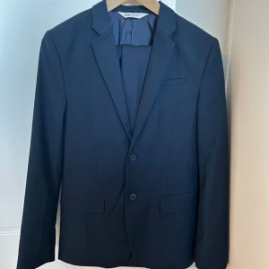 Mörkblå kostym från H&M - Säljer en stilren mörkblå kostym från H&M. Kavajen har två knappar, klassisk krage och raka fickor. Jeansen är raka med pressveck och hällor för bälte. Perfekt för bal eller festliga tillfällen. Den är bara använd 2 gånger sedan den köptes😊