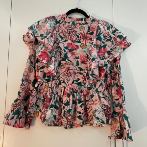En blommig blus - Säljer en blommig rosa blus från Gina Tricot med volangdetaljer runt midjan och vid ämarna samt handleden. Den har en rund halsringning, knappar vid ryggan samt små slits vid ändan av ärmarna. Färgen är mycket ljusare och mer livlig i verkligheten, vid intresse kan skicka fler bilder. Har används ett få antal gånger, men inga synliga tecken på det. Storleken är 38. Priset kan diskuteras. 