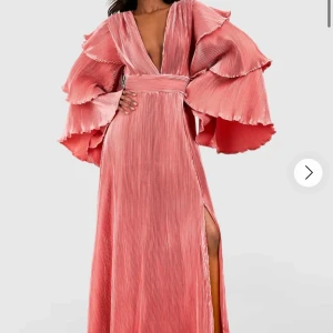 SÖKER DENNA KLÄNNING STORLEK 38/40 ev 36 - Söker en maxiklänning i plisserat tyg med djup v-ringning och breda volangärmar. Klänningen har markerad midja och hög slits framtill. Färgen är en snygg korallrosa som verkligen sticker ut. Klänningen är från boohoo