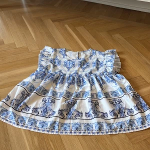 Vit och blå blus med volangärmar - Supersöt vit blus med blått barockinspirerat mönster och volangärmar. Klänningen har rund halsringning och knäppning bak. Perfekt för somriga dagar eller speciella tillfällen.