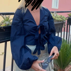 Mörkblå  blus med knyt - Trendig mörkblå blus med croppad passform och knyt framtill. Blusen har långa puffärmar och djup v-ringning. Perfekt för dig som vill sticka ut med en stilren och modern look.