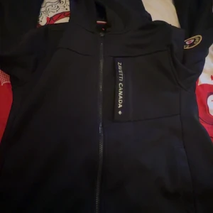 Svart jacka från Zavetti Canada med dragkedja - Säljer en svart jacka från Zavetti Canada med hel dragkedja framtill och huva. Jackan har en diskret lönnlövsdetalj på ryggen och logga på bröstet. Perfekt för dig som gillar stilren design och sportig känsla.