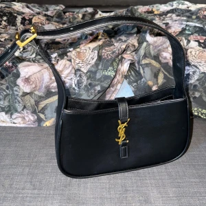 Svart axelväska från Saint Laurent - Snygg svart axelväska från Saint Laurent med guldigt YSL-spänne framtill. Väskan har en stilren, rektangulär form och justerbart axelband med gulddetaljer. Perfekt för att lyfta vilken outfit som helst.