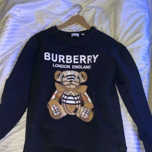 Svart sweatshirt från Burberry med nallebrodyr - Svart sweatshirt från Burberry med stor broderad nalle på framsidan och texten 'BURBERRY LONDON ENGLAND' ovanför. Tröjan har rund halsringning och långa ärmar. Tillverkad i mjuk bomull med elastan för extra komfort.