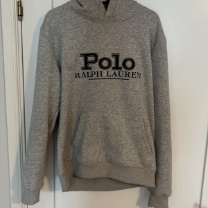 Grå hoodie från Polo Ralph Lauren - Säljer en klassisk grå hoodie från Polo Ralph Lauren med svart logga tryckt på bröstet. Tröjan har huva, lång ärm och en stor magficka framtill. Perfekt för en avslappnad och stilren look. Nästan helt ny använd 3 gånger säljer för att den är för liten. DM för mer bilder eller info✉️ snören tillkommer 