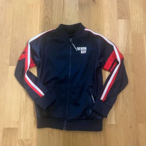 Svart sportig jacka från Denim Day - Säljer en svart sportjacka från Denim Day med röda och vita ränder längs ärmarna. Jackan har dragkedja framtill, ribbade muddar och tryckt logga på bröstet. Perfekt för en aktiv stil.