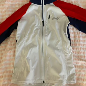 Swix träningsjacka i rött, vitt och blått - Sportig träningsjacka från Swix i vitt med röda och mörkblå detaljer. Jackan har hel dragkedja framtill, hög krage och en bröstficka med dragkedja. Perfekt för utomhusaktiviteter och träning.