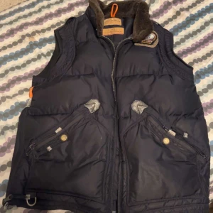 Mörkblå dunväst från Parajumpers - Snygg mörkblå dunväst från Parajumpers med dragkedja och flera fickor framtill. Västen har en hög krage med mjukt foder och detaljer i orange. Perfekt för lager-på-lager under kyliga dagar. Det är en gammal model på parajumper västen vilket gör den mer sällsynt 🤩