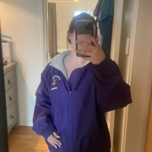 Lila vindjacka med huva - Säljer en lila vindjacka med grå huva och dragkedja framtill. Jackan har tryckt logga på bröstet och är oversized i modellen. Perfekt för kyliga dagar när du vill ha en sportig look.