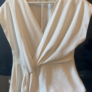 Vit omlottopp med knytband - Säljer en stilren vit omlottopp med knyt i midjan. Toppen har v-ringning och är ärmlös, vilket ger en elegant och modern look. Perfekt att matcha med både jeans och kjol för en trendig outfit.