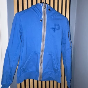 Blå hoodiejacka med dragkedja från Pelle P - Säljer en blå hoodiejacka från Pelle P med grå dragkedja framtill och broderad logga på bröstet. Jackan har huva med snören och fickor på sidorna samt en extra ficka på ärmen.