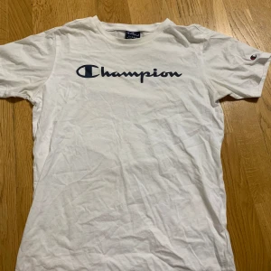 Vit t-shirt från Champion - Vit t-shirt från Champion med klassisk logga tryckt i svart över bröstet. T-shirten har rund halsringning och korta ärmar. Perfekt för en avslappnad och sportig stil.