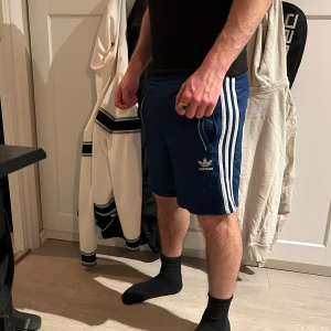 Blå Adidas shorts med vita ränder - Snygga blå shorts från Adidas med klassiska vita ränder längs sidorna och Adidas-logga framtill. Modellen har normal passform och resår i midjan. Perfekta för träning eller en avslappnad stil.