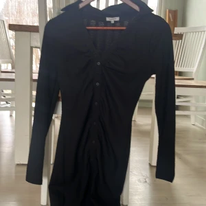 Svart långärmad klänning med knappar - Elegant svart långärmad klänning med knappar framtill. Perfekt för en stilren look. Klänningen har en klassisk krage och en figurnära passform.
