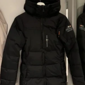 Svart dunjacka från Sail racing - Säljer en svart dunjacka från sail racing. Jackan har en hög krage och dragkedja framtill. Perfekt för kalla dagar med sin vadderade design. Den har långa ärmar och en stilren look.