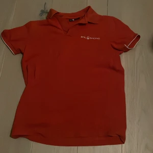 Röd pikétröja från Sail Racing - Snygg röd pikétröja från Sail Racing med vit logga på bröstet. Tröjan har en klassisk krage och korta ärmar med vita detaljer. Perfekt för en sportig look!