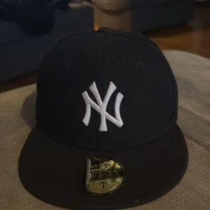 Svart keps från New Era - Snygg svart keps från New Era med broderad vit logotyp på framsidan. Kepsen är en 59FIFTY-modell och har en platt skärm. Perfekt för att ge din outfit en sportig touch.