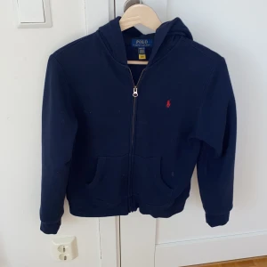 Mörkblå hoodie från Polo Ralph Lauren - Säljer en snygg mörkblå hoodie från Polo Ralph Lauren med dragkedja och huva. Den har en klassisk design med det ikoniska röda logotypen på bröstet. Perfekt för en avslappnad stil. 
