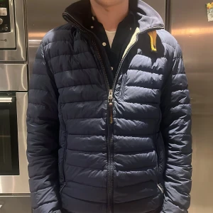 Blå pufferjacka Parajumpers  - Säljer en snygg blå pufferjacka från Parajumpers.(Äkta!) Jackan har en dragkedja framtill och en cool patch på ärmen. Perfekt för kyligare dagar med sin quiltade design och lätta material. 