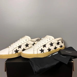 Vita sneakers med svarta stjärnor från Saint Laurent - Snygga vita sneakers från Saint Laurent med svarta stjärnor på sidorna. Skorna har en klassisk snörning och en beige sula. Perfekta för en stilren look med en touch av rock'n'roll.