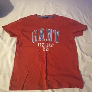 Röd t-shirt från GANT - Säljer en röd t-shirt från GANT med trycket 'EAST COAST 1949' på framsidan. Den har en klassisk rund hals och korta ärmar. Perfekt för en avslappnad stil.