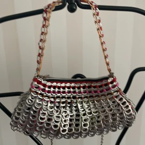 Unik handväska av metallringar - Säljer en unik handväska/axelrem gjord av återvunna metallringar / läsköppnare med färgglada detaljer i rött, rosa och orange.Mått 25*15 cm. Väskan har en dragkedja upptill och en kedja som axelrem. Perfekt för dig som vill ha något annorlunda och miljövänligt!